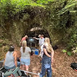 Wide view of a film crew working in front of the ivy-covered stone entrance arch of the cave, beneath the woodland canopy
