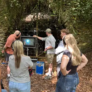 Film crew in headphones gathered around a monitor in front of the ivy-covered gated entrance of the cave