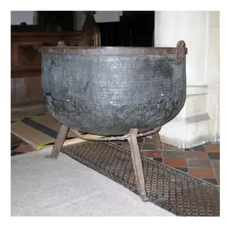 An old, large, black metal cauldron or pot with a rounded shape, sitting on three metal legs, placed indoors on a tiled floor with a rug nearby.