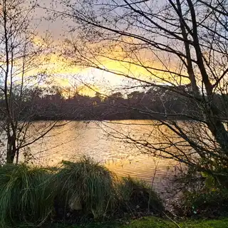 A serene lakeside at sunset with leafless trees in the foreground and a house with a deck on the left. The sky is colorful with hues of orange, purple, and blue, and the free-flowing water reflects the sky's colors.