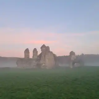 Ruins of an ancient stone castle surrounded by green grassy field and mist, with a pink and blue sky at dawn or dusk in the background.